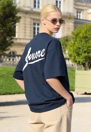 Femme blonde portant des lunettes de soleil marron, chemise bleu marine avec un texte blanc "Ignore" dans le dos, et pantalon beige, à l'extérieur près de buissons verts et d’un bâtiment.