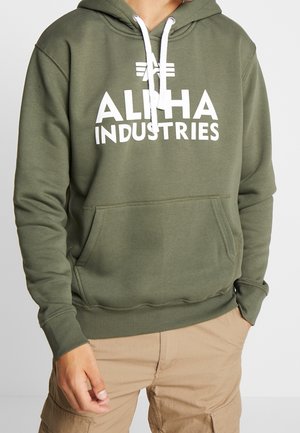 Person iført en olivengrøn Alpha Industries hættetrøje med hvide snore og khaki cargobukser, hænderne i lommerne.