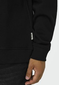 Zwarte sweatshirt met geribbelde boorden en zoom, met een label aan de zijkant. Soepele textuur. Hand zichtbaar tegen donkere jeans.