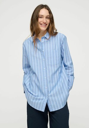 Smilende kvinde med langt brunt hår iført en løs lyseblå skjorte med striber og mørke bukser, hænderne i lommerne, foran en hvid baggrund.