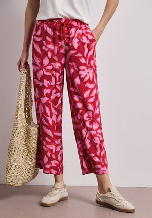 Kobieta w szerokich spodniach w czerwono-różowe kwiaty, białych tenisówkach, białej bluzce, trzyma beżową szydełkowaną torbę.