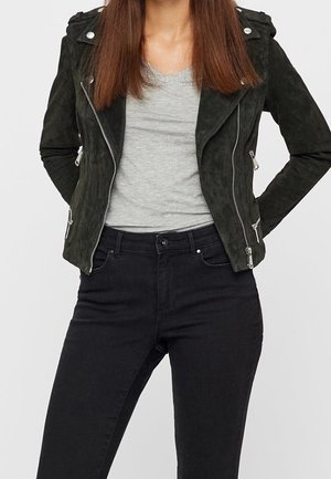 Femme portant une veste de motard en daim noire avec des fermetures éclair argentées, un t-shirt gris clair et un jean noir taille haute, debout les mains derrière le dos.