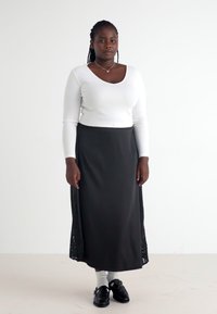 Hvit ribbestrikket langermet topp og svart skjørt med blonder på sidene. Svarte sko og hvite sokker. Enkelt bakgrunn.