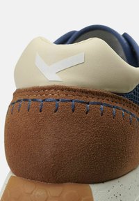 Dettaglio di sneaker con tacco in pelle beige, texture in suede marrone, cuciture blu e accentuazione in bianco del logo. Suola in gomma esagonale visibile.