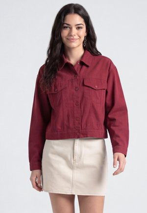 Jeune femme aux cheveux foncés portant une veste bordeaux boutonnée et une jupe beige clair, souriante devant un fond clair uni.