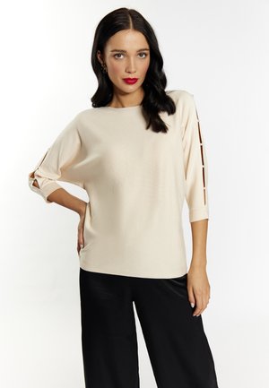 Cremfarbener Pullover mit Dreiviertelärmeln, ausgestattet mit Aussparungen an den Ärmeln und einer strukturierten Stoffoberfläche. Kombiniert mit einer schwarzen Hose.