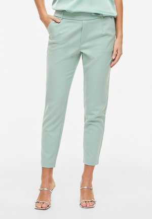 Kvinde iført lysegrønne smalle bukser og sølvfarvede, stropper høje hæle, den ene hånd i lommen, mod hvid baggrund.