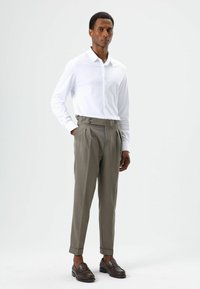Chemise blanche boutonnée à manches longues associée à un pantalon gris taille haute avec plis et deux boutons latéraux. Des mocassins marron complètent la tenue.