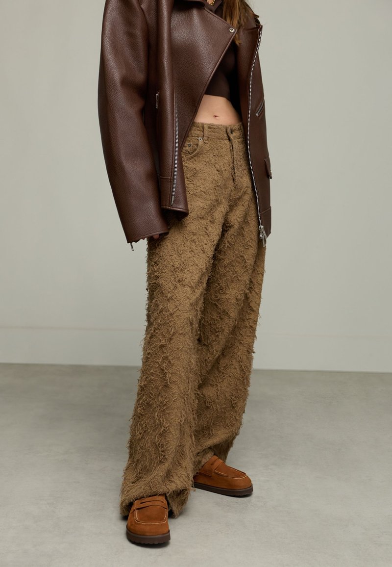 Personne portant un pantalon large texturé marron, un top court marron, un manteau en cuir oversized marron foncé et des mocassins en suede marron se tenant sur un sol gris.