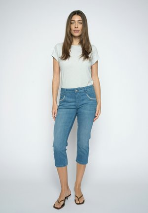 Vrouw staand, draagt een licht gestreept T-shirt, blauwe korte jeans en bruine sandalen met hak tegen een effen lichte achtergrond.