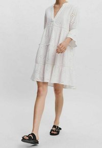 Vit, knälång klänning med V-ringning, trekvartsärmar, ögla detaljer och lager på lager volanger vid nederkanten. Bärs med svarta sandaler.