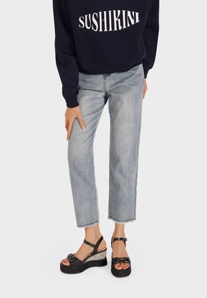 Persona che indossa jeans corti azzurri chiaro e sandali con zeppa neri con cinturini alla caviglia, con la mano appoggiata sulla coscia.