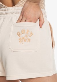 Beige Baumwollshorts mit einem elastischen Bund und einer Gesäßtasche, die mit einem gestickten 'ROXY'- und Blattdesign versehen ist, betont die weiche Textur.
