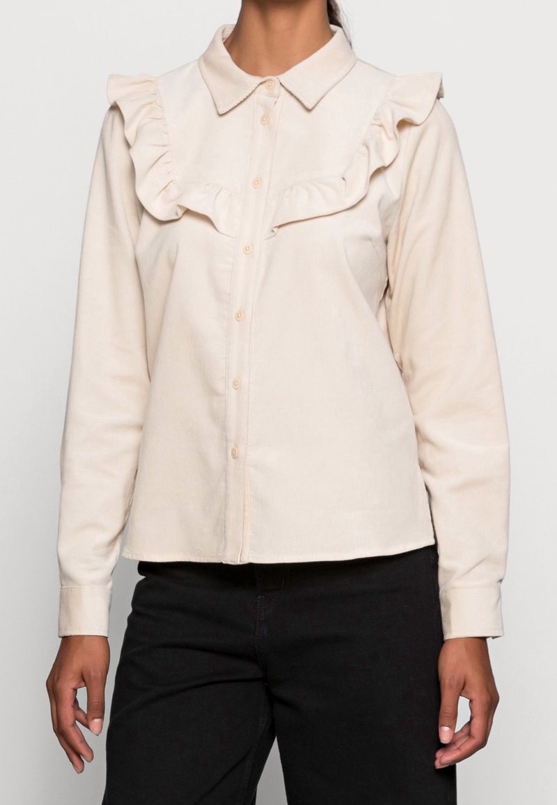 Cream button-up shirt with a ruffled yoke, long sleeves, and a collar. Made of a smooth, lightweight fabric. Paired with black pants.