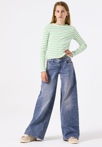 Groene en witte gestreepte longsleeve top gecombineerd met brede blauwe denim jeans en beige sneakers, staande tegen een effen achtergrond.