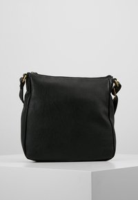 Svart läder crossbody-väska med guldfärgad metallhardware och justerbar axelrem, vilande på en vit pelare.