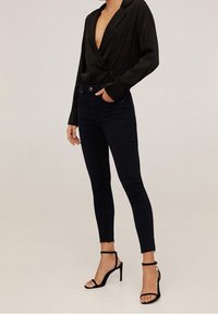 Mujer vestida con blusa negra de escote en V profundo, jeans skinny negros con dobladillos deshilachados y sandalias de tacón alto con tira al tobillo negras, de pie.