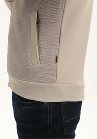 Beżowy sweter z teksturowaną siateczką, boczną kieszenią na zamek błyskawiczny i ściągaczem u dołu. Połączone materiały oferują kontrast w projekcie i teksturze.