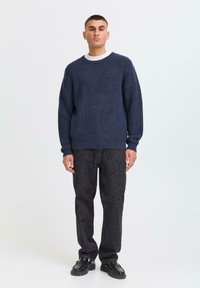 Pull en maille marine à finition texturée, avec un col rond large et des poignets côtelés, associé à un pantalon en denim noir et des chaussures noires brillantes.