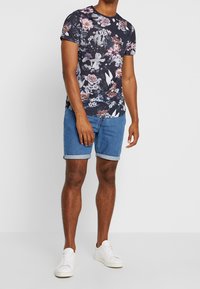 La camiseta floral presenta un fondo oscuro con flores rosas y blancas. Combinada con pantalones cortos de denim y zapatillas blancas. Diseño de mangas cortas.