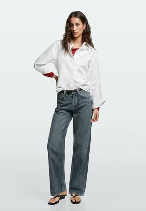 Femme aux longs cheveux foncés portant une chemise blanche boutonnée sur un haut rouge, un jean sombre ample et des tongs noires, debout devant un fond uni.