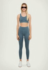 Dopasowany zestaw odzieży sportowej w stonowanym odcieniu teal. Biustonosz z okrągłym dekoltem, legginsy z wysokim stanem, oba wykonane z gładkiego, elastycznego materiału. Białe sneakersy.