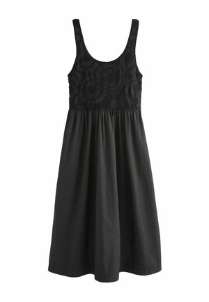 Vrouw met lang blond haar, die een mouwloos zwart gehaakt top draagt, een zwarte maxi rok, een zwarte riem, gelaagde kettingen en zwarte sandalen.