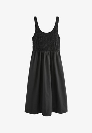 Kobieta z długimi blond włosami ubrana w czarny, szydełkowany top bez rękawów, czarną maksisukienkę, czarny pasek, warstwowe naszyjniki i czarne sandały.