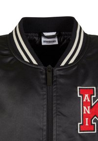 Černá bomber bunda s bílým proužkovaným žebrovaným límcem, předním zipem a červenou varsity nášivkou s písmeny "A N I K" na hrudi.