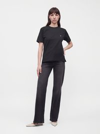 Femme debout, une main sur la hanche, portant un t-shirt noir, un jean gris foncé et des chaussures claires à bout pointu sur un fond uni.
