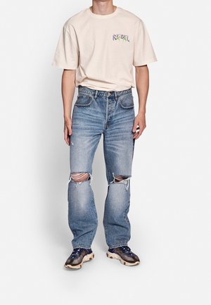 Homme portant un t-shirt beige à manches courtes avec un texte graphique et un jean bleu clair déchiré aux genoux, assorti à des baskets bleues et grises.