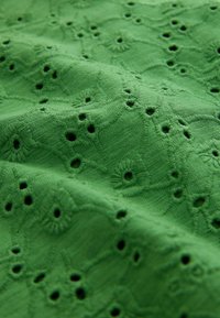 Tissu vert avec broderie florale en relief et motifs de découpes circulaires régulièrement espacés, créant une surface texturée et décorative.