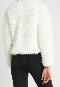 Hvid, fluffy jakke med en blød, tekstureret finish. Har en rund kant og lange ærmer, parret med mørk denimjeans.