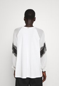 Camisa blanca de manga larga con detalles de encaje negro en las mangas, de corte holgado y puños fruncidos; etiqueta en el cuello.