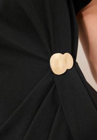 Must mustriga, pehmest kangast, millel on voldid ja kuldne ümmargune detail, mis kinnitab kogutud materjali, luues struktureeritud aktsendi.