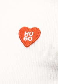 Un tissu blanc côtelé présente une écusson en forme de cœur orange avec un texte blanc indiquant "HUGO". La broderie ajoute de la texture et du détail.