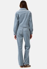 Frau in einer hellblauen Cordjacke und passenden Hosen, mit dem Rücken zur Kamera stehend, trägt braune und weiße Sneaker.