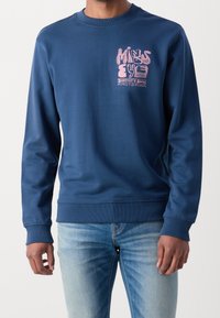 Granatowa bluza z bawełnianej mieszanki, z okrągłym dekoltem, z różowym nadrukiem na piersi. Ściągacze na mankietach i u dołu. Zestawiona z jasnoniebieskimi dżinsami.