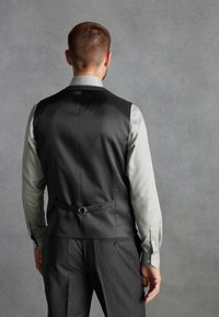 Vesta neagră cu o finisare netedă, cu hardware pe curea în spate, asortată cu o cămașă gri deschis și pantaloni gri cu textură. Detalii subtile de croi vizibile.