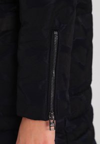 Manga de chaqueta de plumas negra con un patrón de camuflaje, abertura con cremallera y herrajes metálicos, textura suave y diseño acolchado.