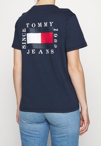 Laivastonsininen puuvillainen t-paita, jossa on selässä rohkea kuvio, punavalkoinen suorakaiteen muotoinen logo sekä tekstit "TOMMY JEANS" ja "1985".