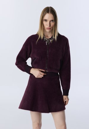 Paarse nep-bont zip-up jas met geribbelde manchetten, gecombineerd met een bijpassende flared rok. Sieraden met sleutels worden om de hals gedragen.