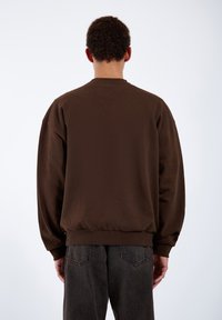 Felpa oversize marrone con scollatura a girocollo, polsini e orlo a coste, realizzata in un materiale morbido, abbinata a jeans in denim scuro.