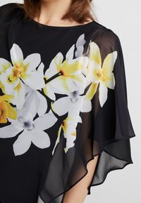 Haut transparent noir avec imprimé floral présentant de grandes fleurs blanches et jaunes, design fluide et manches volantées. Texture lisse et forme drapée.