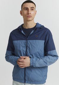 Giacca leggera blu con design a blocchi di colore contrastanti in navy, chiusura con zip, cappuccio regolabile e polsini elastici. Texture liscia.