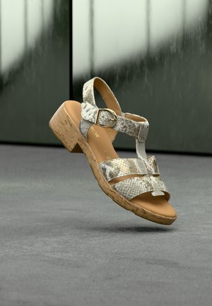 Sandale pour femme à motif peau de serpent beige et gris, avec un petit talon en liège et une boucle dorée, flottant au-dessus d'un sol gris avec un arrière-plan sombre flou.