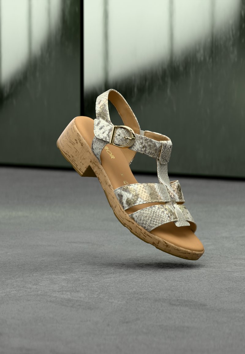 Sandale pour femme à motif peau de serpent beige et gris, avec un petit talon en liège et une boucle dorée, flottant au-dessus d'un sol gris avec un arrière-plan sombre flou.