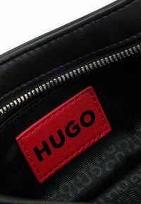 Interior de cuero negro con un zipper plateado. Presenta una destacada etiqueta de cuero rojo con "HUGO" estampado en negro. Fondo sutilmente texturizado.