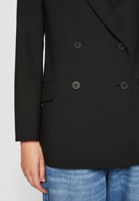 Blazer nero doppiopetto in tessuto testurizzato, con quattro bottoni neri, rever a scendere e tasca laterale, abbinato a jeans azzurri chiari.