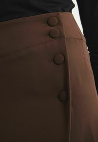 Jupe marron en tissu lisse avec un design ajusté. Dispose de quatre grands boutons ronds sur un côté et d'un ourlet net.